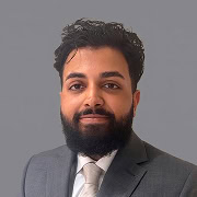 A man with dark hair and a beard, wearing a gray suit, white shirt, and light-colored tie, poses in front of a plain gray background.