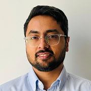 A man with short black hair, a beard, and glasses is wearing a light blue collared shirt and standing in front of a plain white background.