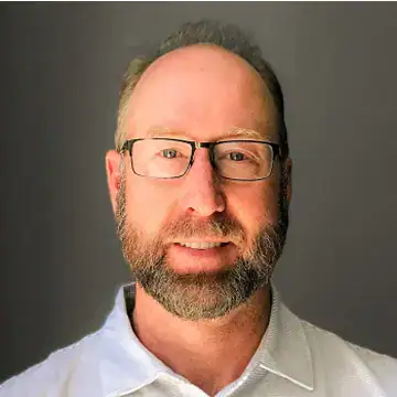 A man with glasses, short light brown hair, and a trimmed beard smiles at the camera. He is wearing a white collared shirt and is set against a plain, dark background.