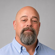 Bald man with a gray beard and mustache, wearing a light blue collared shirt, posing in front of a plain gray background.