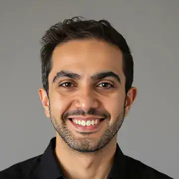 A man with short dark hair and a trimmed beard smiles at the camera. He is wearing a black shirt and is posed against a plain gray background.