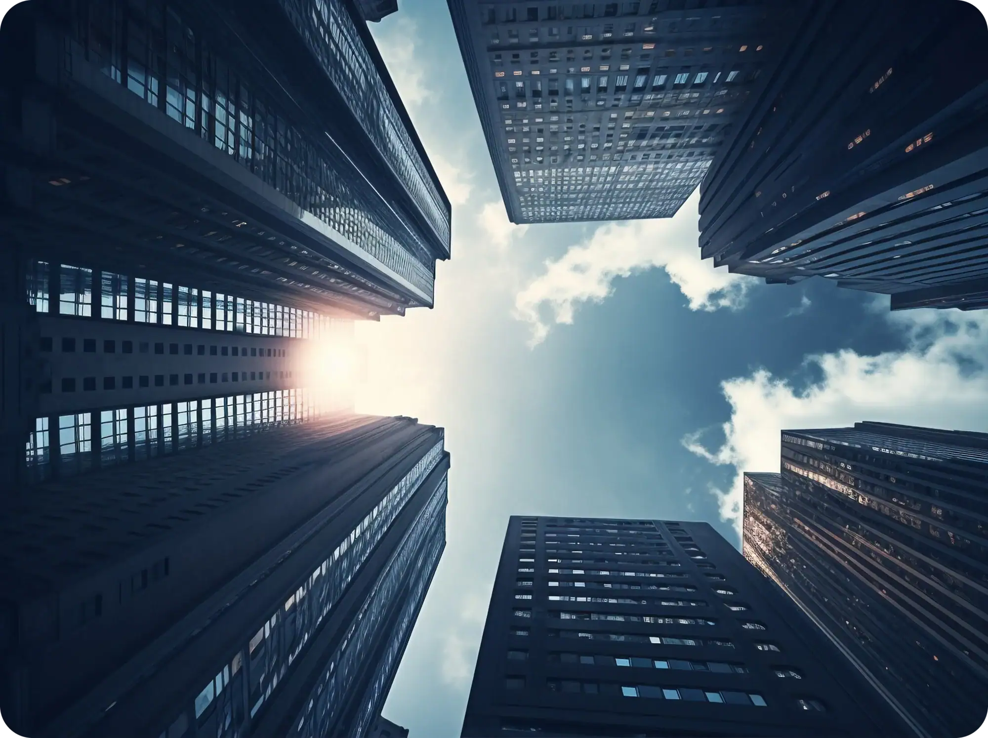 Skyscrapers viewed from below with the sun shining between the buildings. The sky is partly cloudy, creating a dramatic urban cityscape.