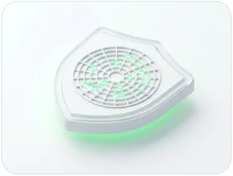 White, shield-shaped electronic device with a circular vent pattern on top and a glowing green light beneath it, placed on a light-colored surface.