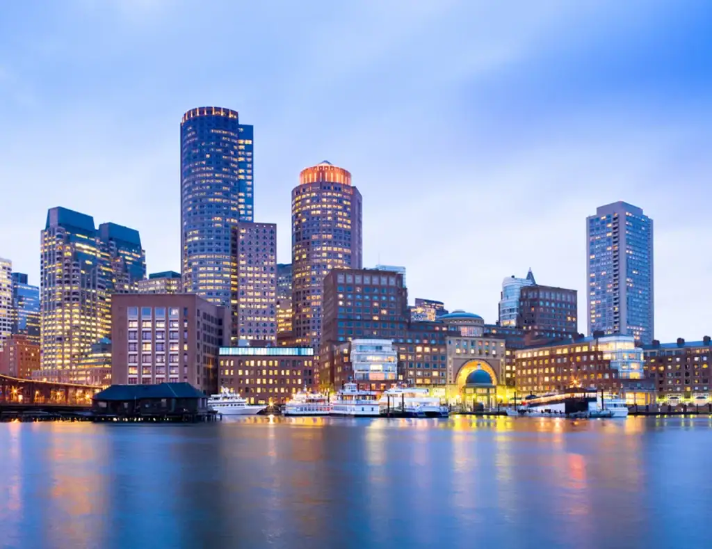Boston city skyline at dusk, with illuminated skyscrapers reflecting on the water and boats docked along the waterfront under a blue sky.