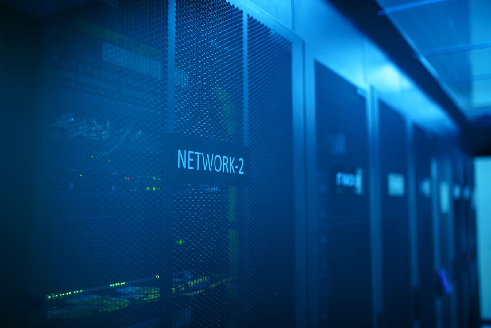 A row of server racks in a data center, illuminated by blue light. One rack is labeled "NETWORK-2" and features visible grilles and faintly glowing indicator lights.