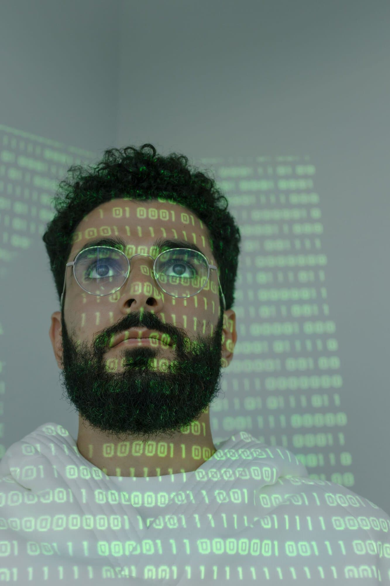 A man with glasses and a beard stands against a gray wall, with green binary code projected onto his face and the background, creating a digital and futuristic atmosphere.