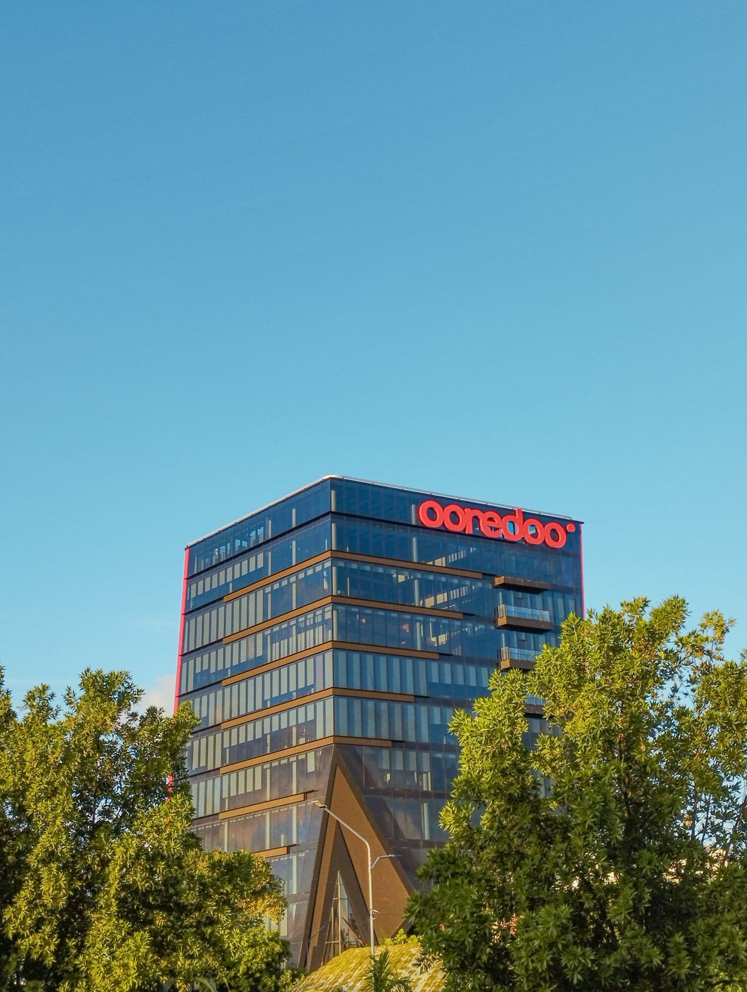 A modern glass office building with a red "ooredoo" sign on top, surrounded by green trees, under a clear blue sky.
