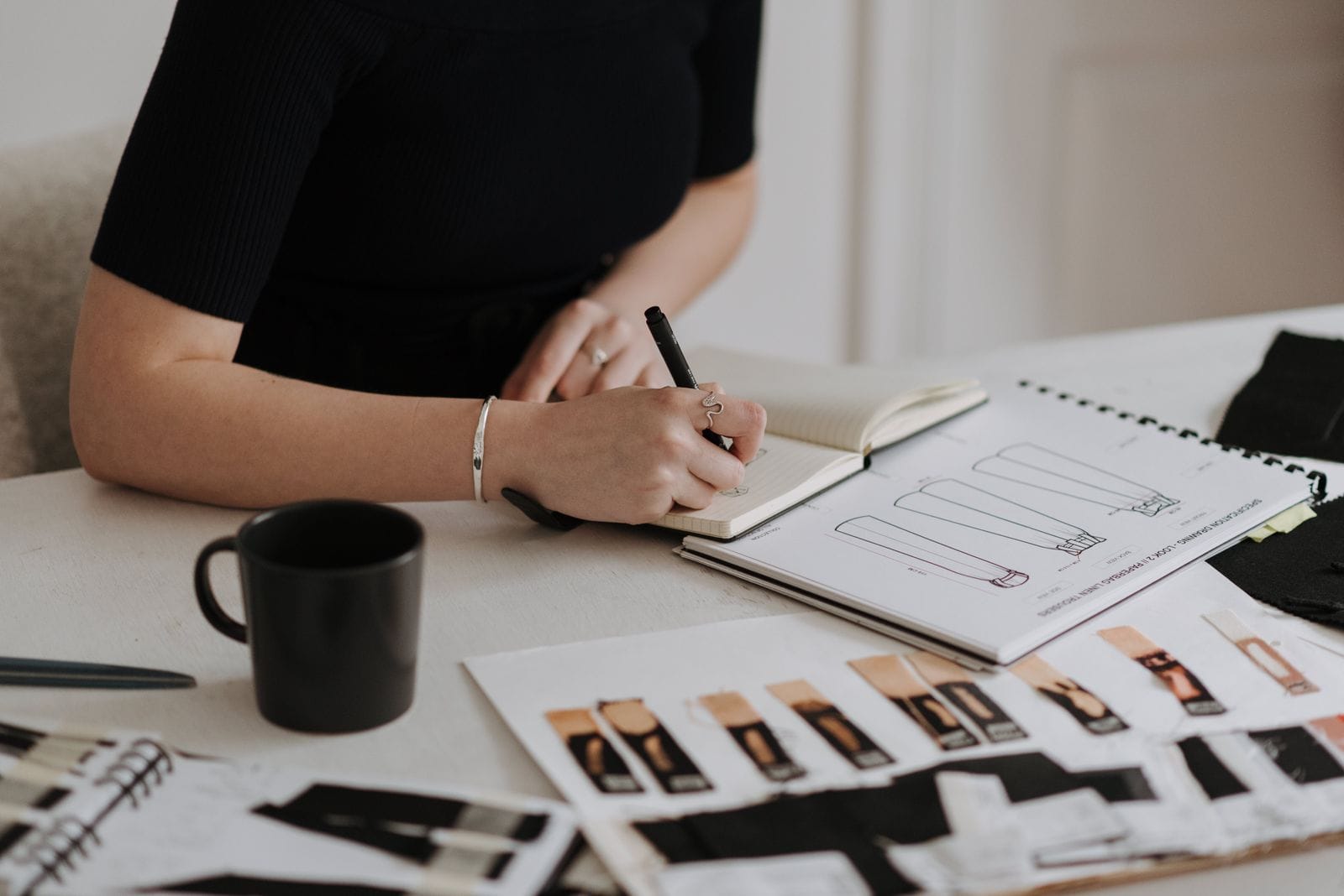 A person writes in a notebook at a desk with a coffee cup, sketches, and fabric samples, suggesting work on fashion or interior design concepts.