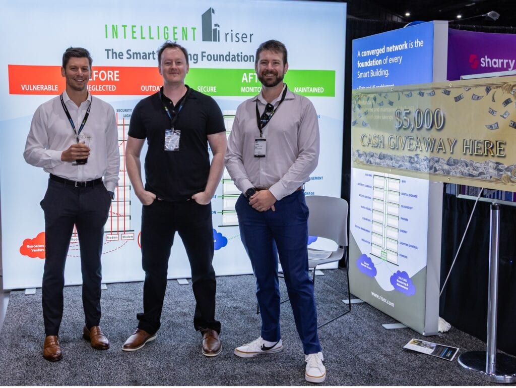 Three men stand smiling in front of a tech booth display with charts and a sign that reads “$5,000 Cash Giveaway Here.” The booth promotes a smart building solution at an indoor event.